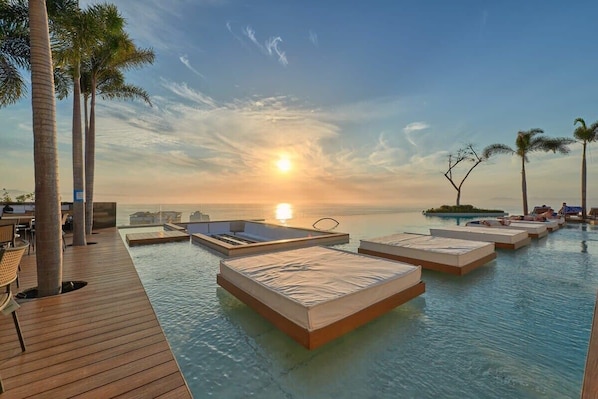 A poolside area with sunbeds and a wooden deck, overlooking a body of water with a sunset in the background.