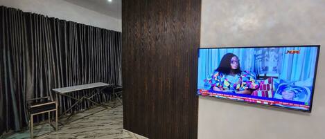 A room with a television mounted on the wall, a table with chairs, and a patterned floor.