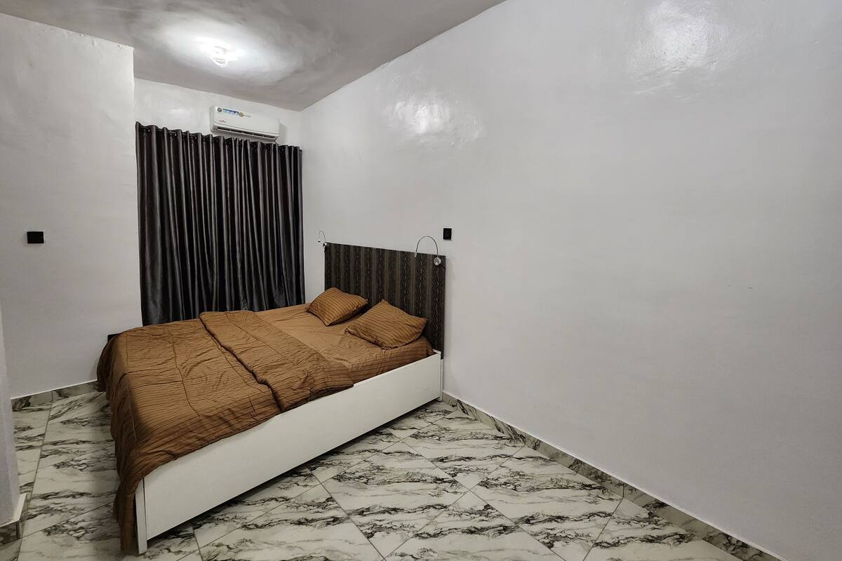 A bedroom with a bed, a headboard, a window with curtains, and a wall-mounted air conditioner.