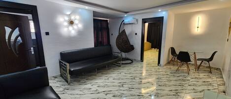 Un salon moderne avec un canapé noir, un fauteuil suspendu, une table basse en verre et un coin repas comprenant des chaises et une table.