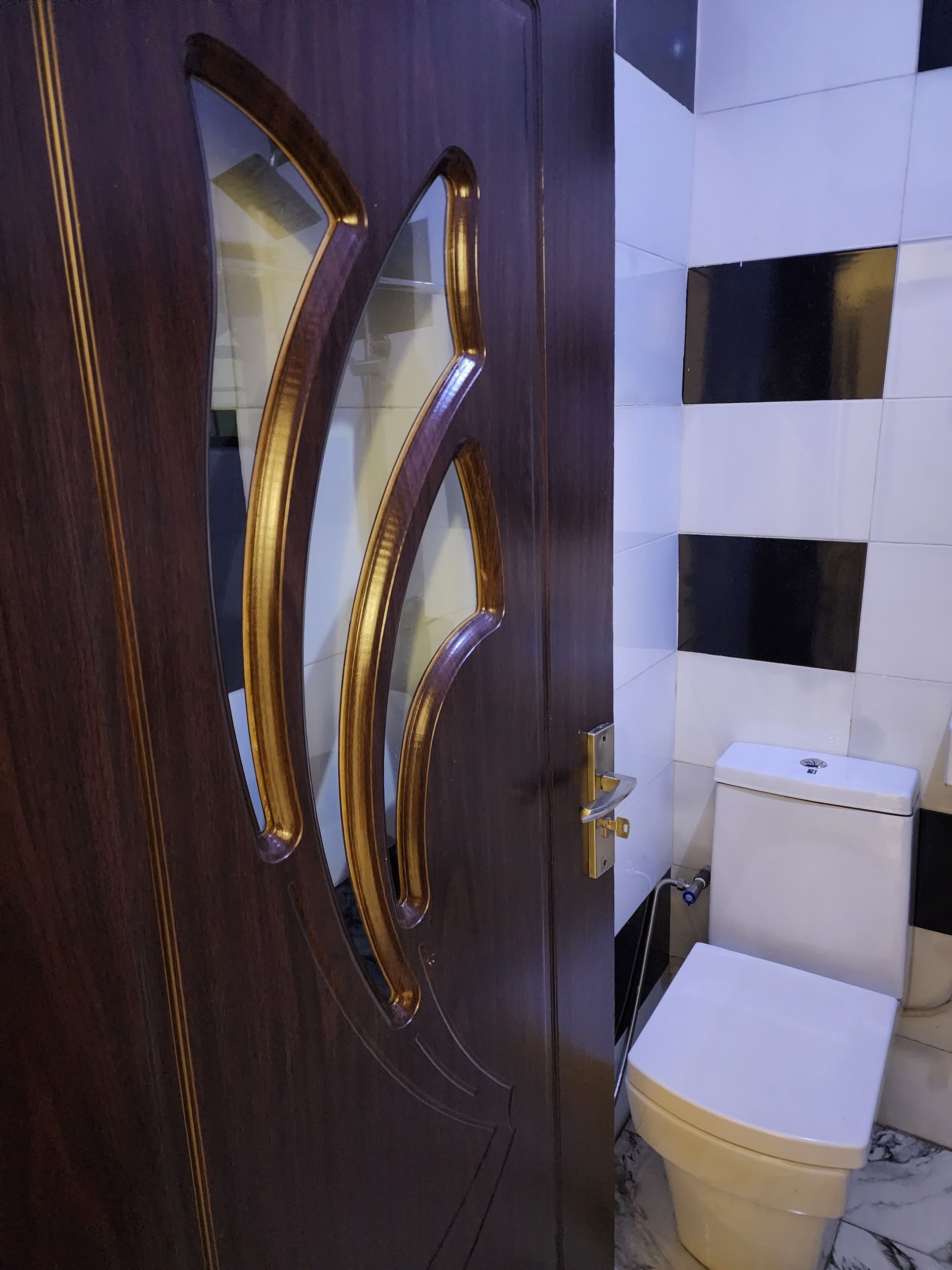 A wooden door with a gold handle and a glass panel, a toilet, and a tiled wall with black and white tiles.