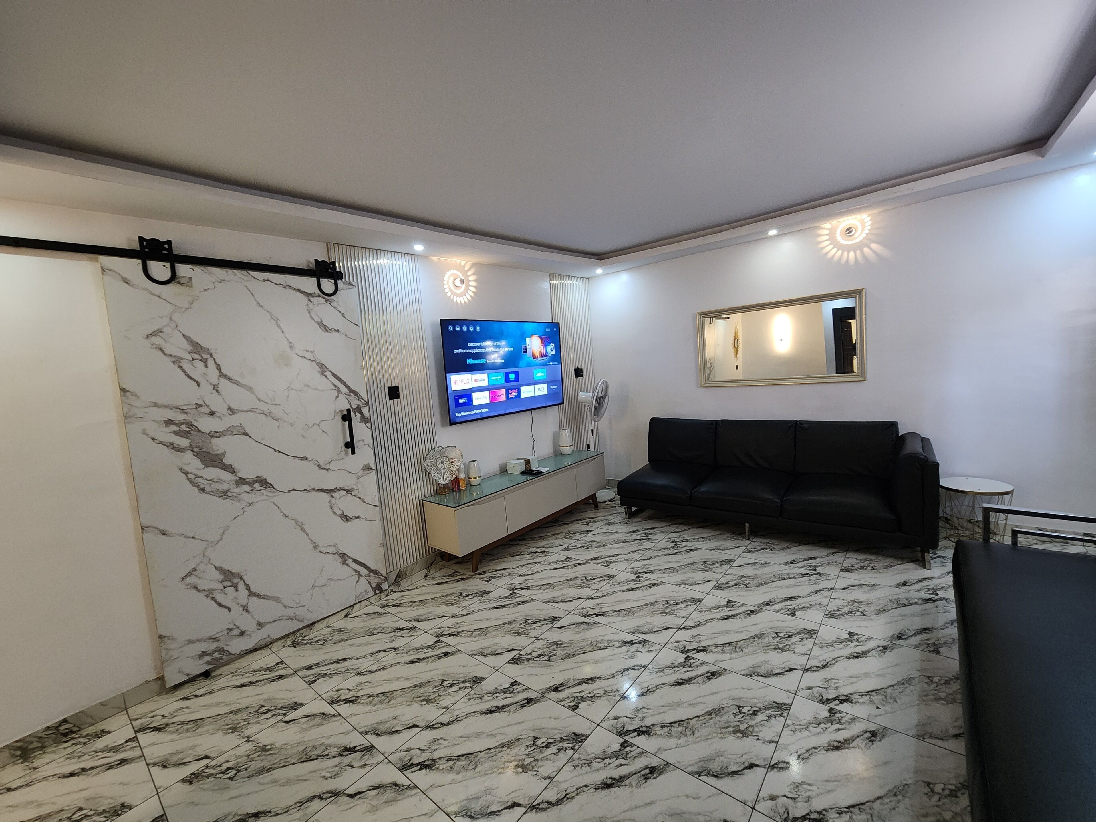 Een moderne woonkamer met een marmeren vloer, een zwarte bank, een aan de muur bevestigde televisie en een klein tafeltje met een ventilator.