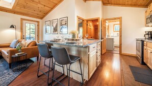 Un intérieur de chalet chaleureux avec un plafond en bois, un coin repas avec un bar, une cuisine et un salon doté d’un canapé et de fauteuils.