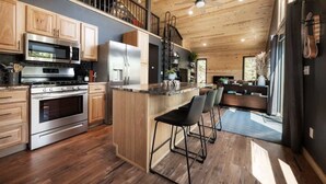 Een moderne keuken met houten kasten, roestvrijstalen apparatuur, een centraal eiland met barkrukken, en een trap naar een verdieping erboven.