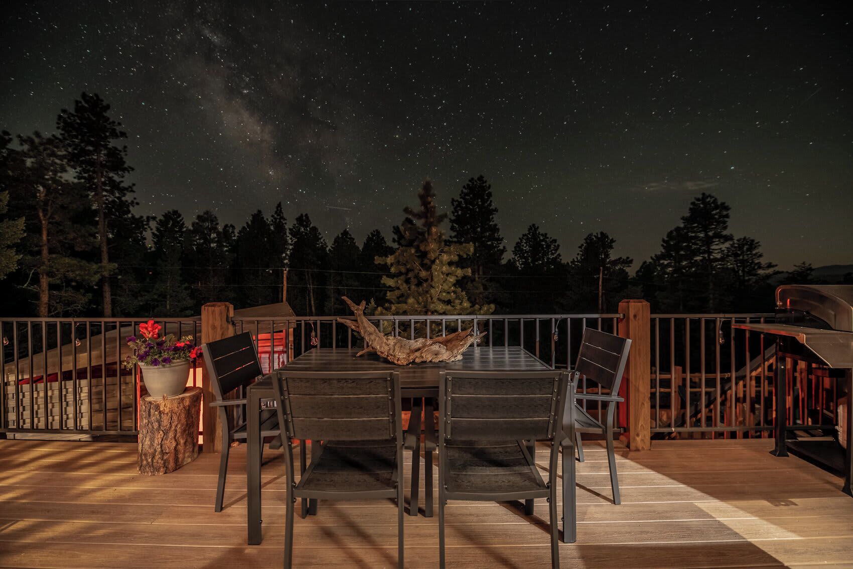 An outdoor dining area with a table and chairs, surrounded by a wooden deck and railing, under a night sky with visible stars.