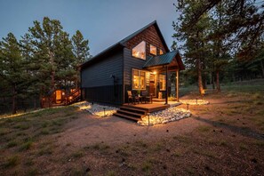 A cabin with a deck, surrounded by trees, and a gravel path leading to the entrance.