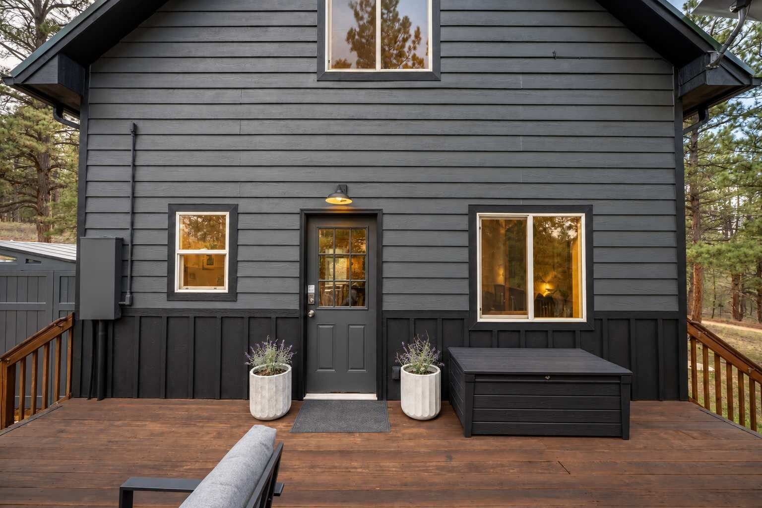 A two-story house with a dark exterior, a front door, two windows, a porch with a bench, and planters.