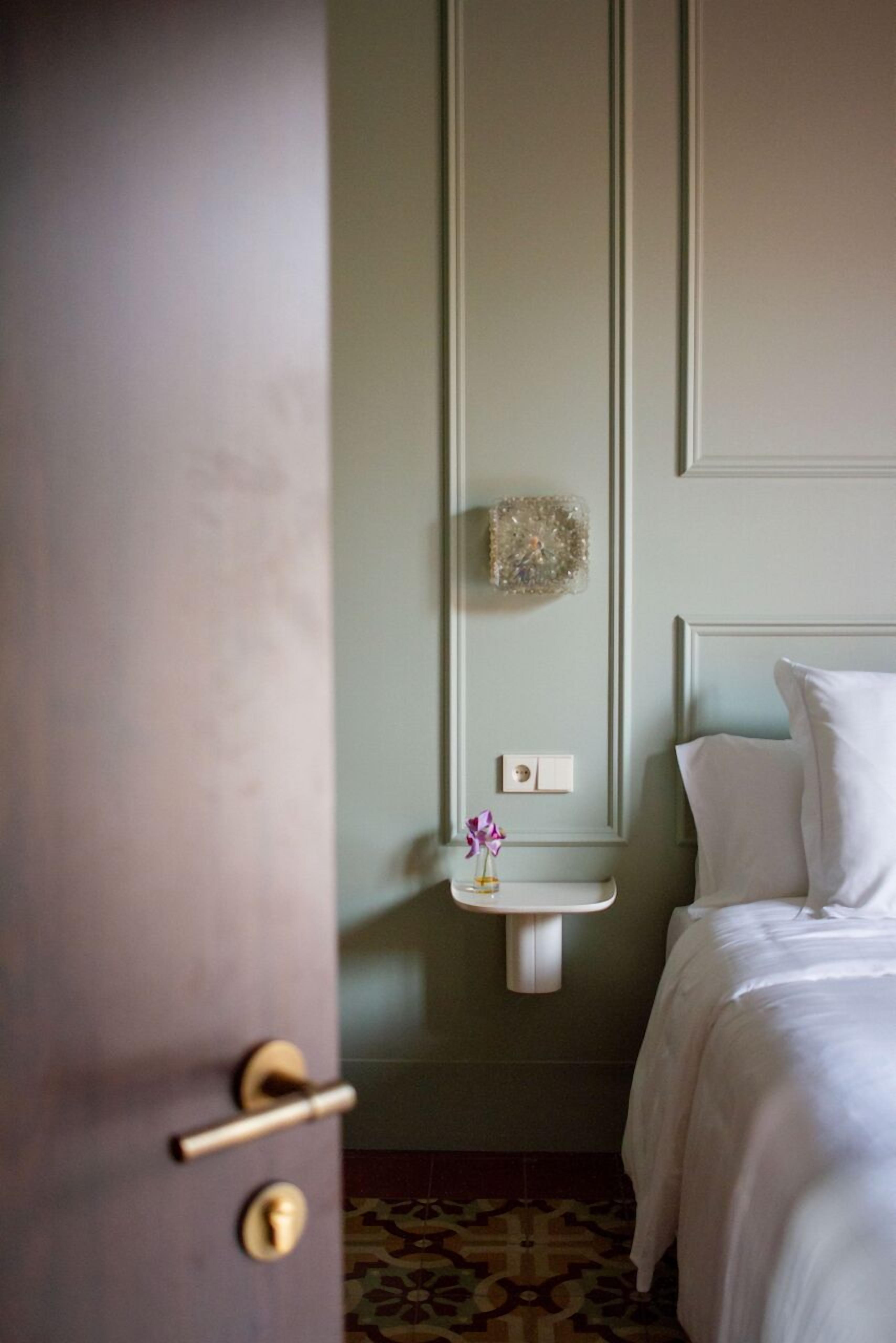 Une chambre d’hôtel avec un lit, une table de chevet ornée d’un vase de fleurs, un luminaire fixé au mur et une porte dotée d’un trou de serrure.