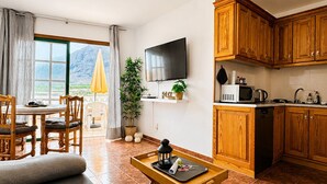 Een woonkamer met een bank, een salontafel, een televisie en een keukenhoek met houten kasten en een magnetron.
