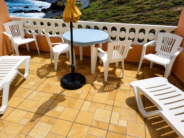 Um terraço com cadeiras de plástico brancas e uma mesa redonda, um guarda-sol amarelo e vista para o oceano.