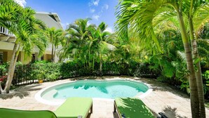 Ein Poolbereich mit einem gewundenen Pool, umgeben von Palmen und Grünpflanzen, sowie zwei grünen Liegestühlen.