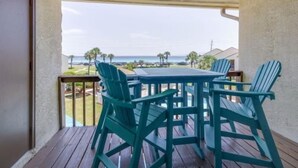 A balcony with a wooden table and chairs, overlooking a beach with palm trees and a clear sky.