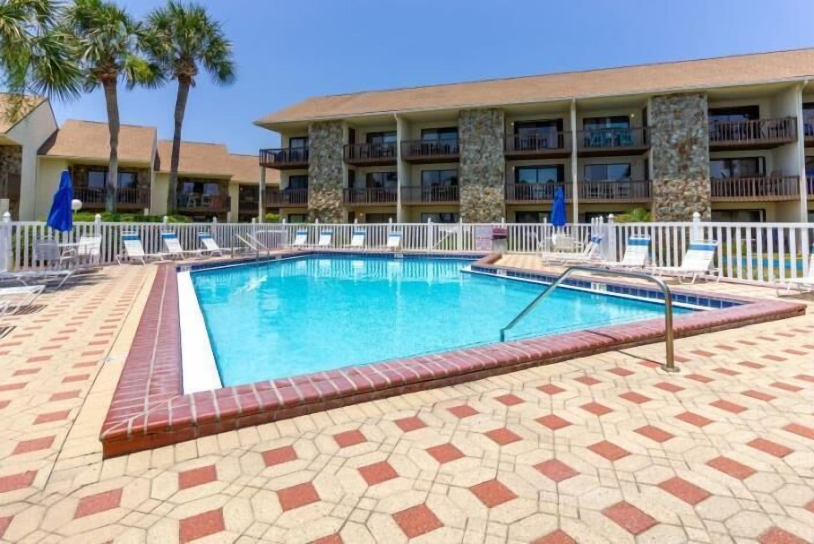 A hotel pool area with a rectangular pool, lounge chairs, and a multi-story building with balconies.