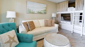 A living room with a teal sofa, a white sofa, and a round ottoman. There is a painting on the wall and a ceiling fan.