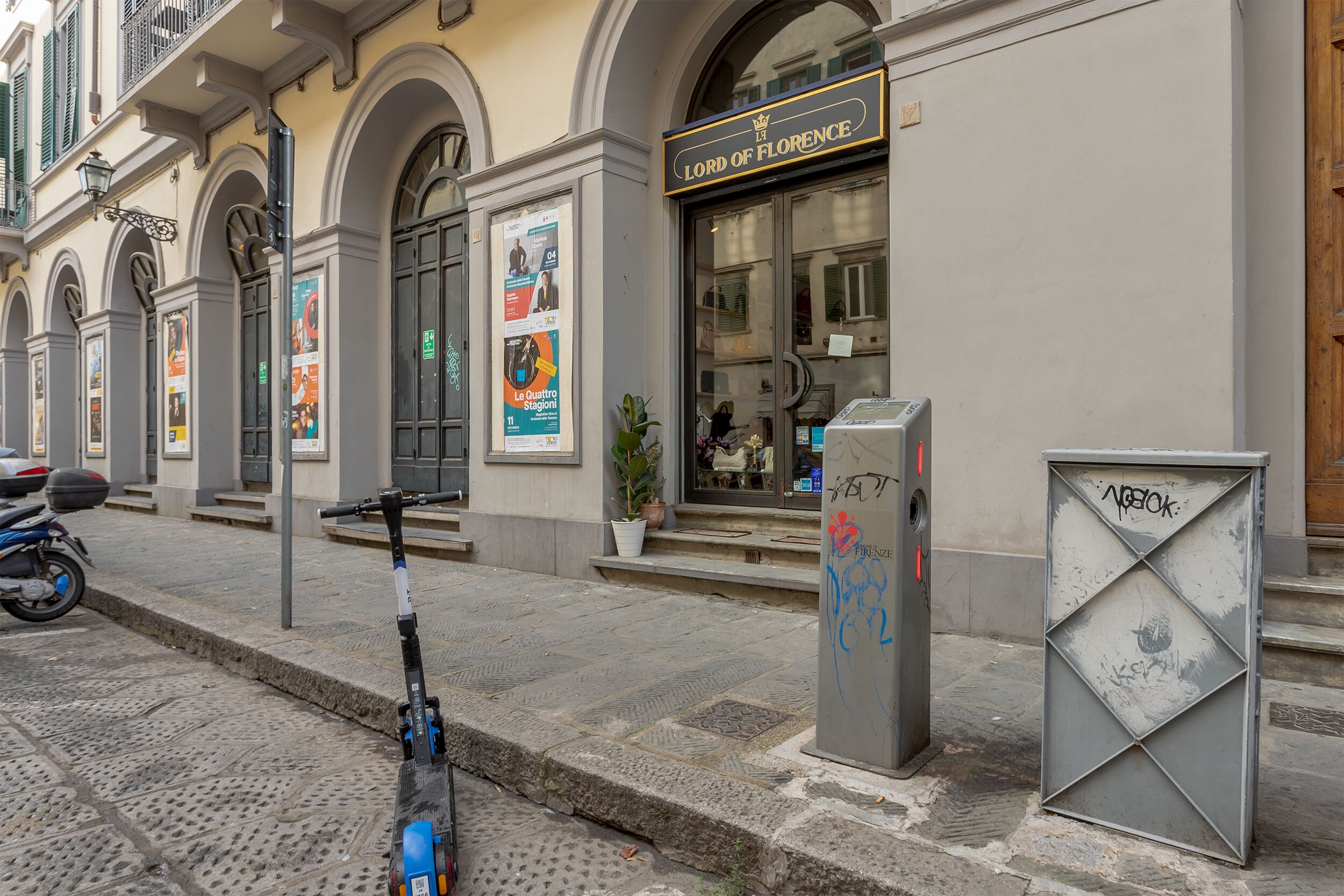 Une rue où un scooter est garé sur le trottoir, un immeuble avec une enseigne indiquant « Lord of Florence », et un coffret électrique orné de graffitis.