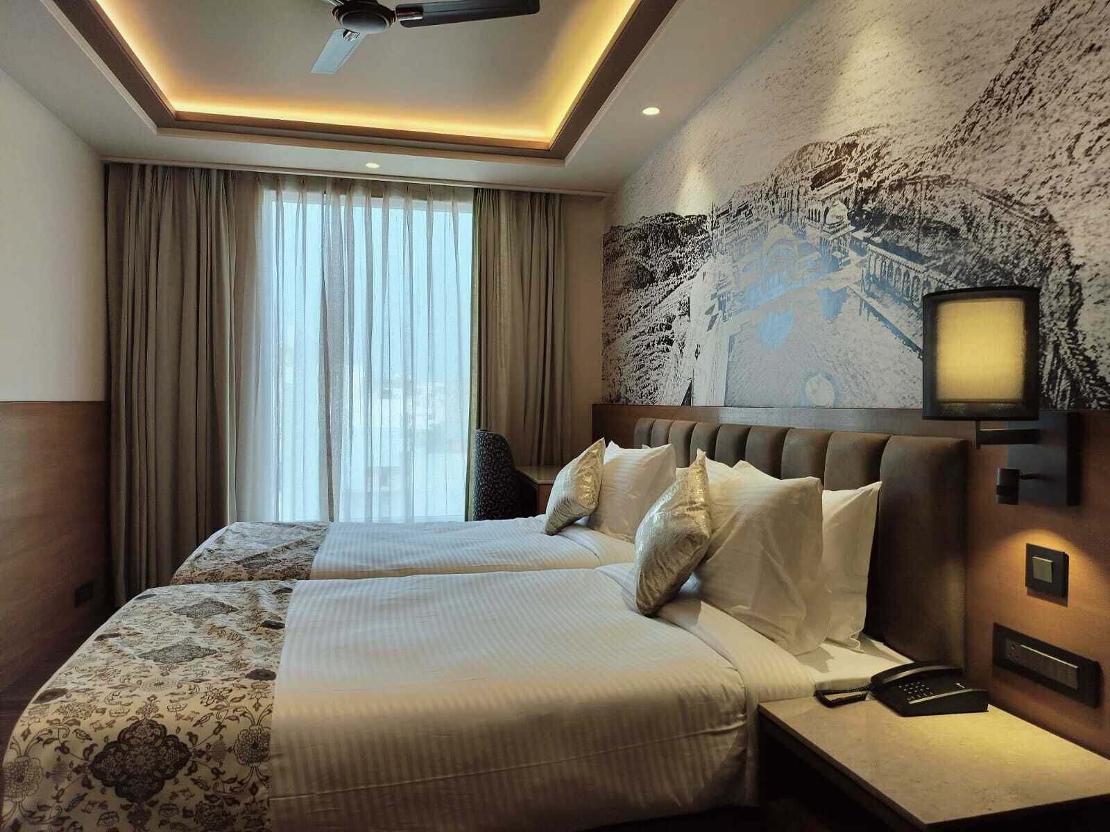 Une chambre d’hôtel avec deux lits, une grande fenêtre avec des rideaux, un ventilateur de plafond, un téléviseur fixé au mur et un téléphone sur la table de chevet.