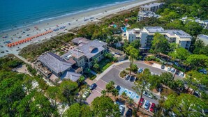 An aerial view of a coastal hotel with a beach view, parking area, and multiple buildings.