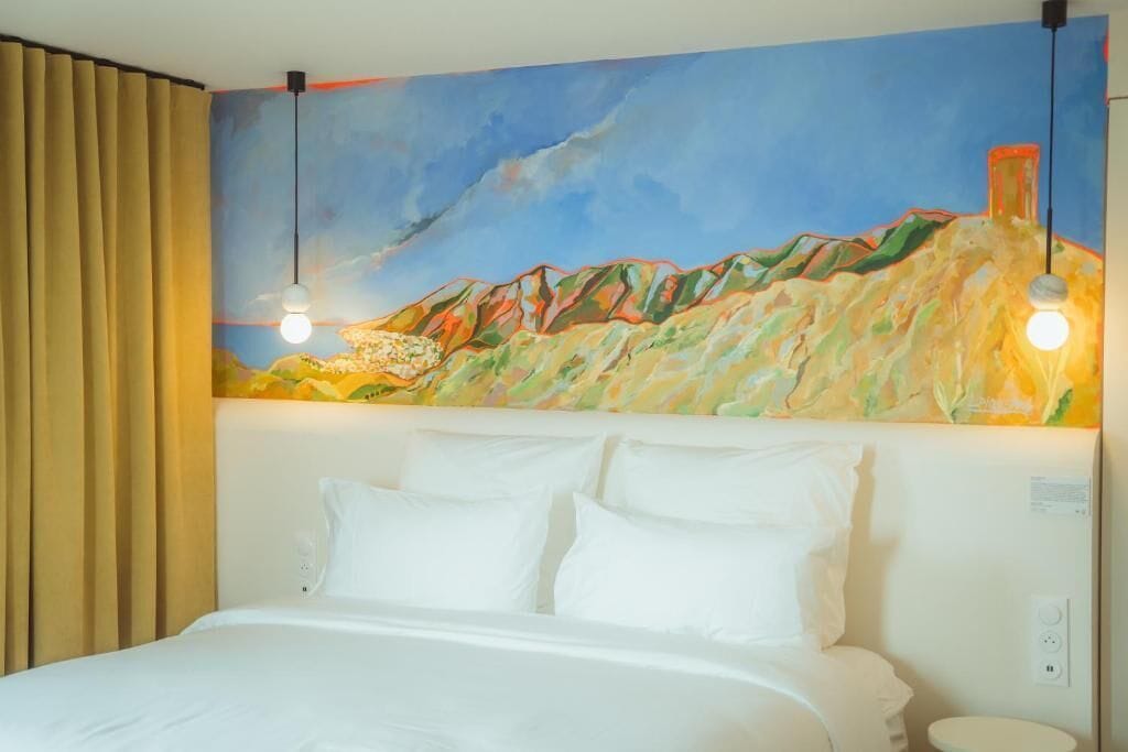 A hotel room with a bed, white pillows, a painting of a mountain landscape on the wall, and two pendant lights.