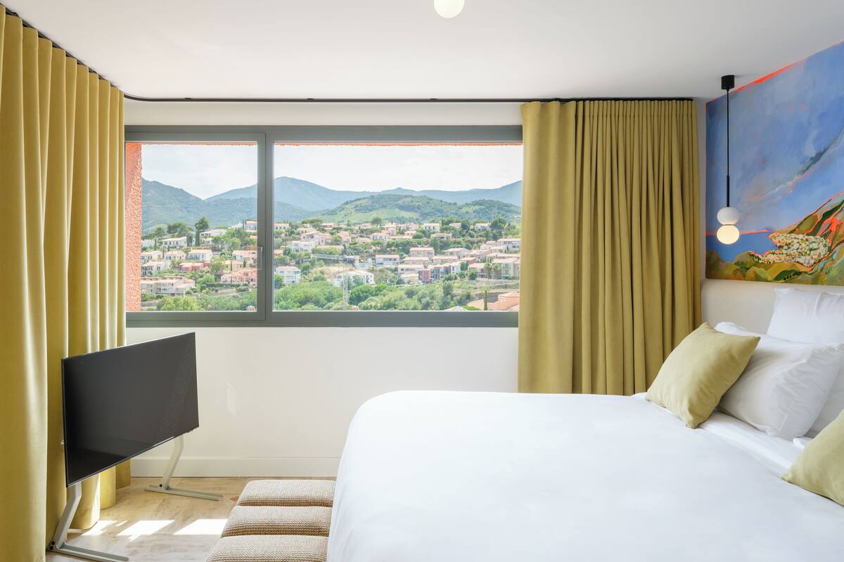Ein Hotelzimmer mit Bett, Fernseher und großem Fenster mit Blick auf eine Stadt und Berge.