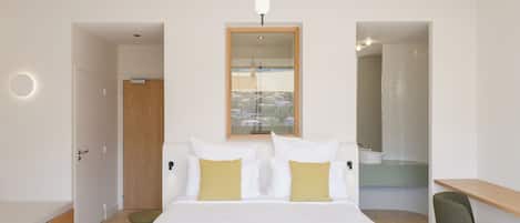 A modern bedroom with a large bed, a desk, and a view of the city through a window.