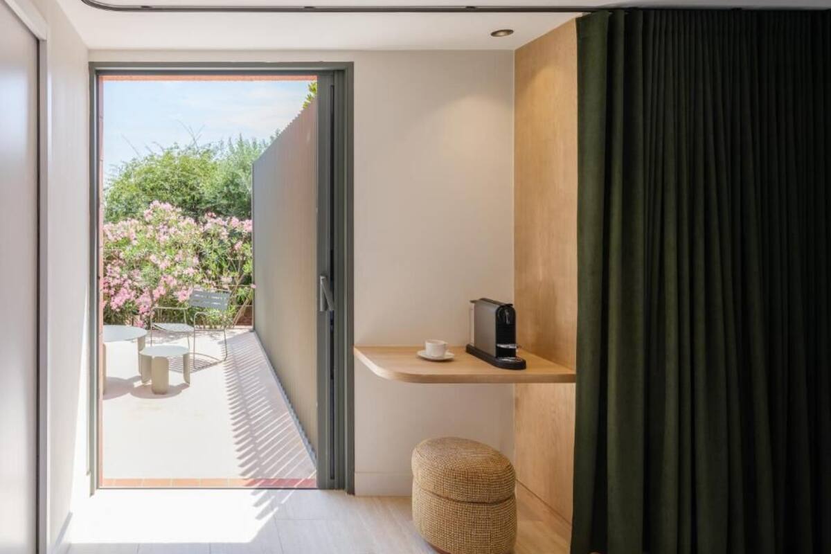 Ein modernes Zimmer mit einer Schiebetür zu einer Außenveranda, auf der ein Tisch und Stühle stehen. Es gibt ein kleines Holzregal mit einer Kaffeemaschine und einen gewebten Hocker.