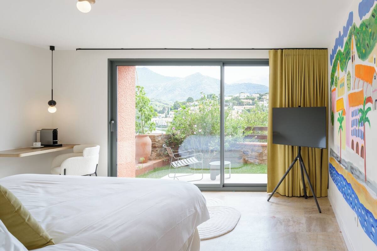 Ein Schlafzimmer mit Bett, Schreibtisch mit Kaffeemaschine, Fernseher auf einem Standfuß und Blick auf Garten und Berge durch eine Schiebetür.