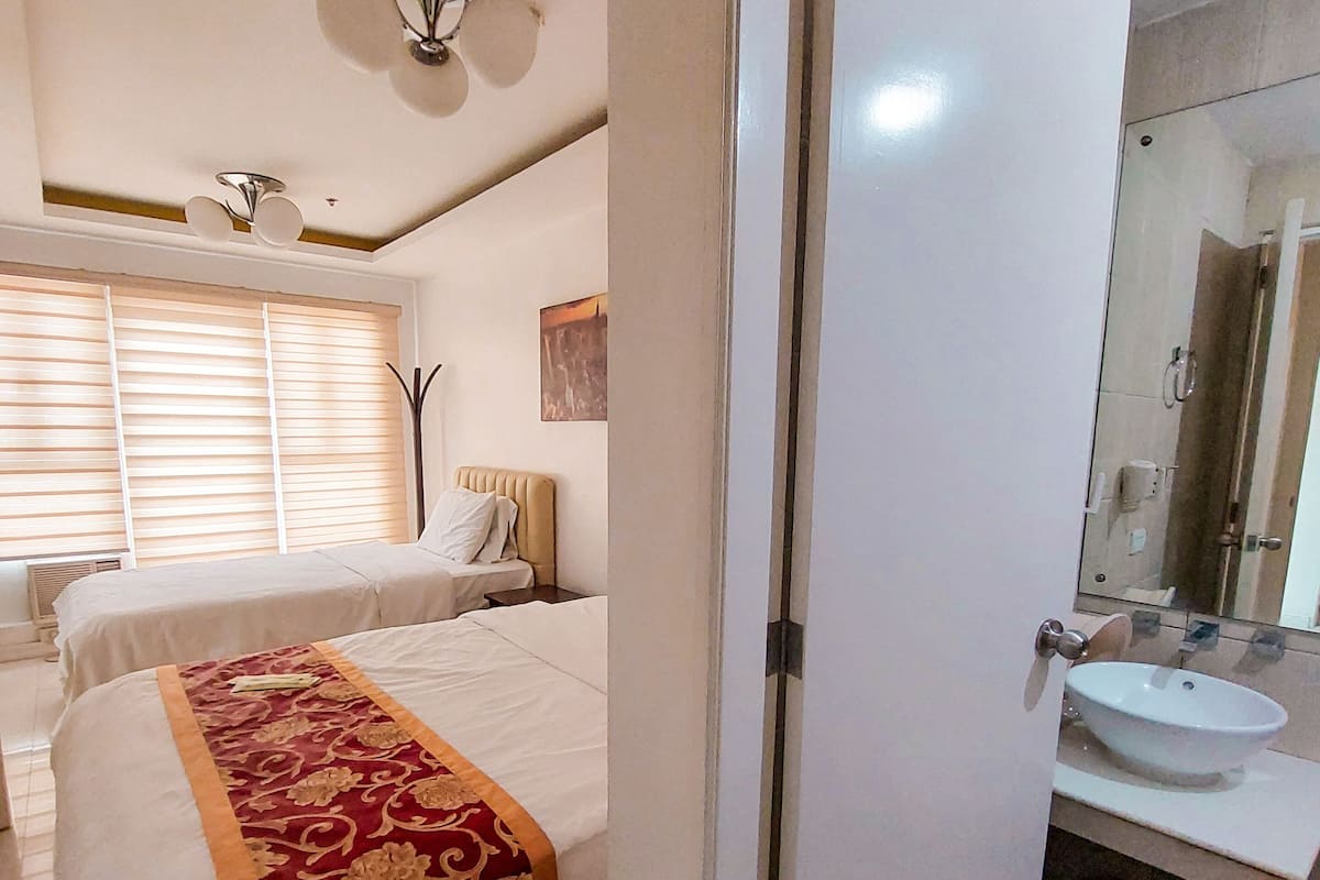 Une chambre d’hôtel comprenant un lit, une table de chevet, une fenêtre avec des stores, un ventilateur de plafond et une salle de bain avec un lavabo et un miroir.