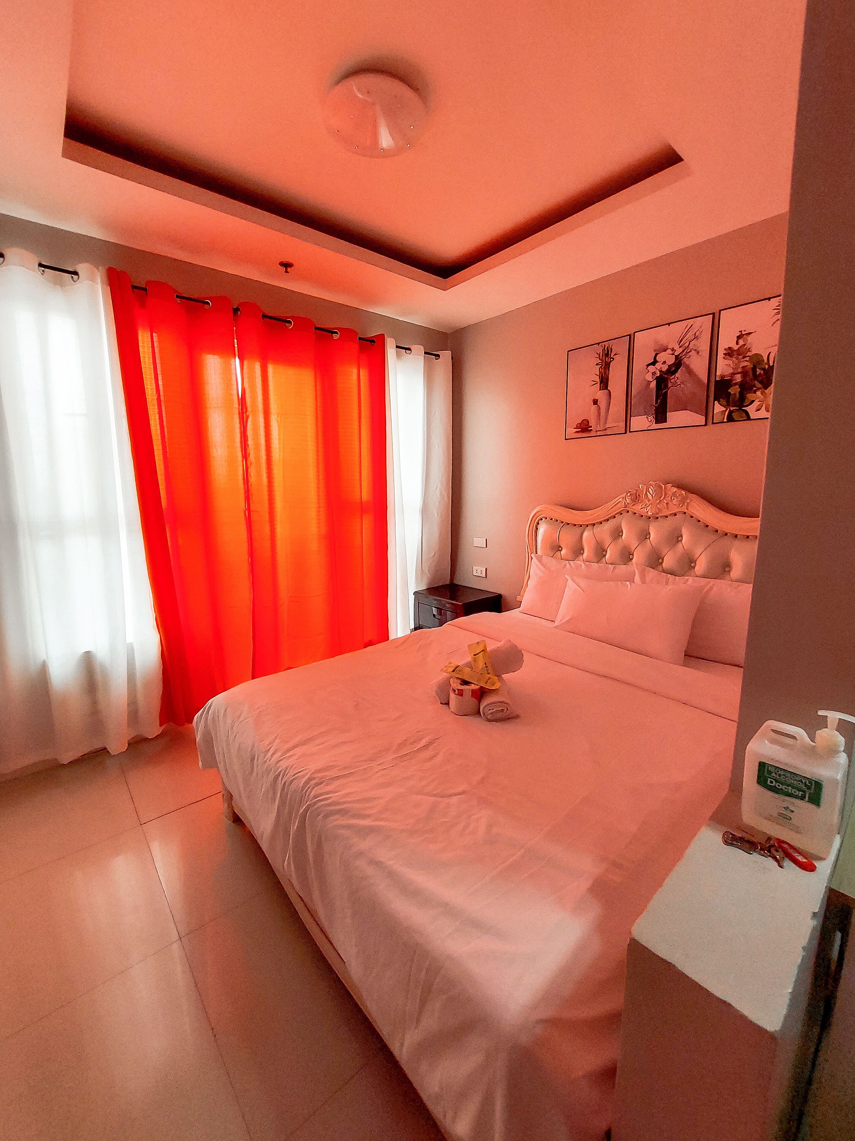 A hotel room with a large bed, a nightstand, a wall-mounted clock, and a window with red curtains.