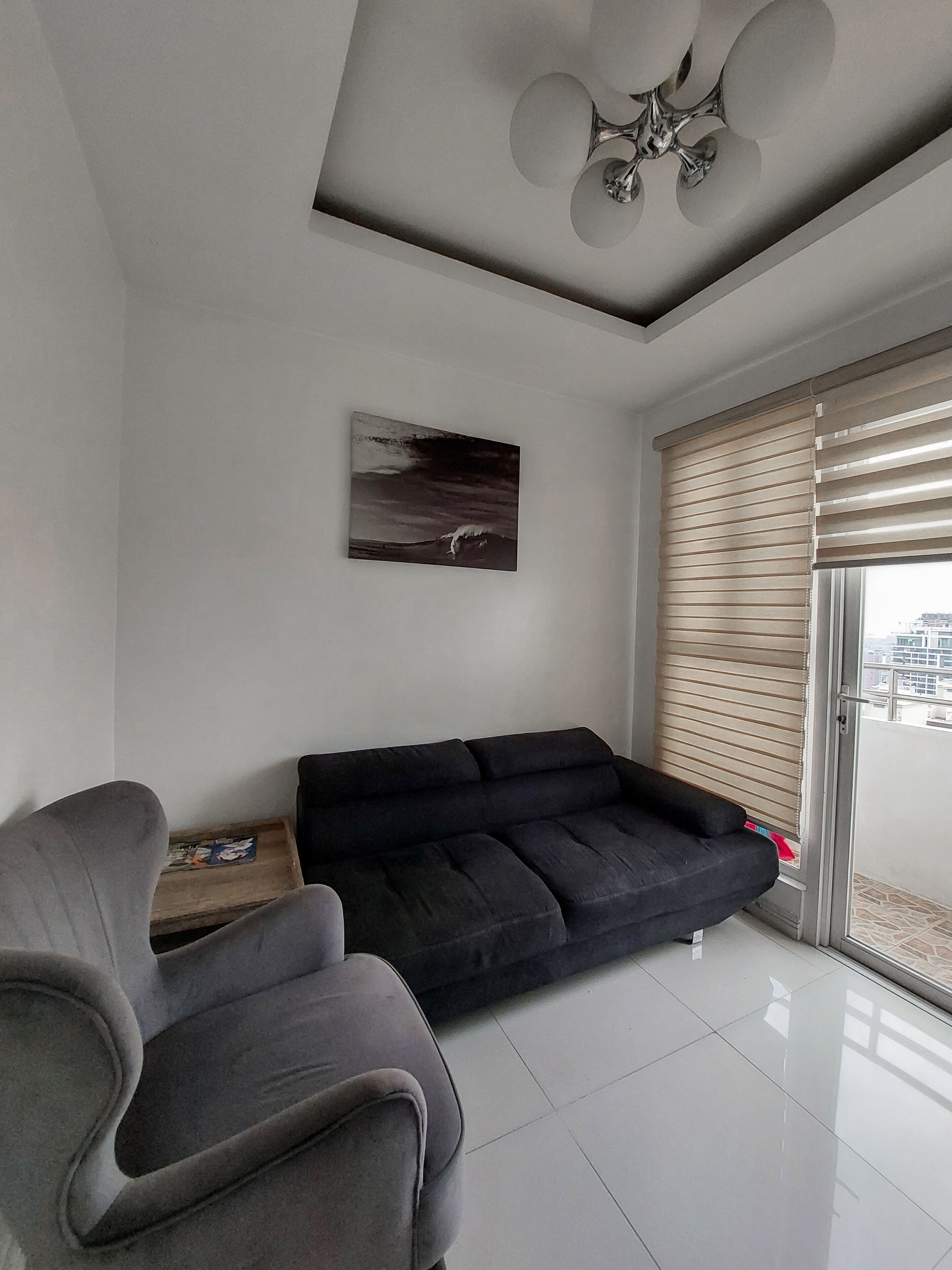 A modern living room with a black sofa, a grey armchair, a ceiling fan, a painting of a wave, and a large window with blinds.