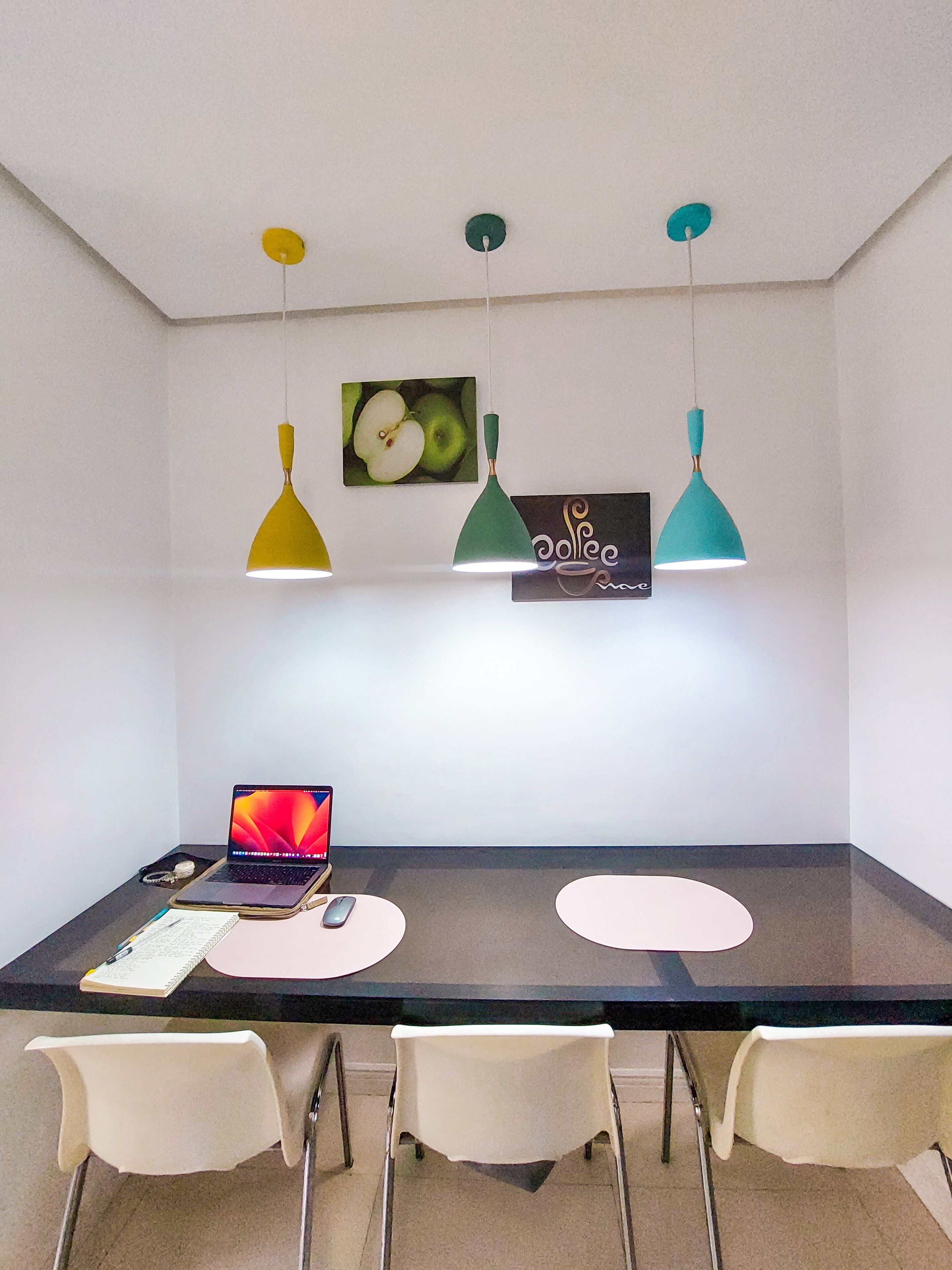 Un comedor moderno con una mesa oscura, tres lámparas colgantes y un ordenador portátil sobre la mesa.