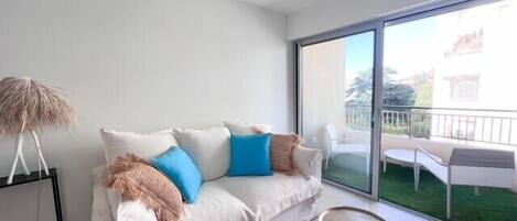 A modern living room with a white sofa, blue and beige cushions, a small round table, and a floor lamp. A large sliding glass door leads to an outdoor area with a patio table and chairs.