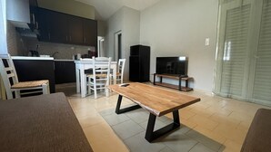 A modern living room with a wooden coffee table, a TV on a stand, and a dining area with white chairs and a kitchen in the background. - Milo by the sea (Analipsi)