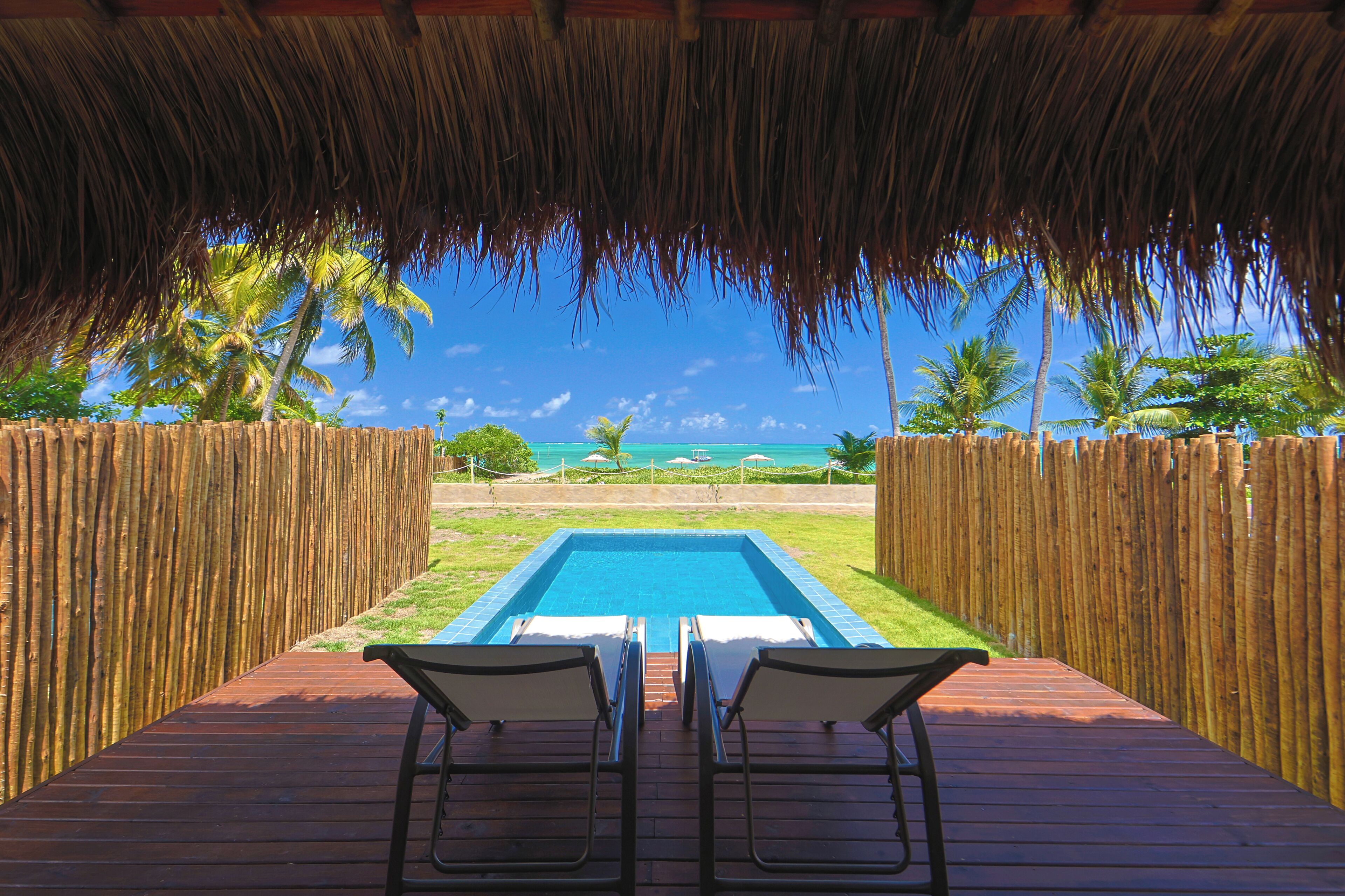 A wooden deck with a pool and two lounge chairs facing a clear blue sky and palm trees.