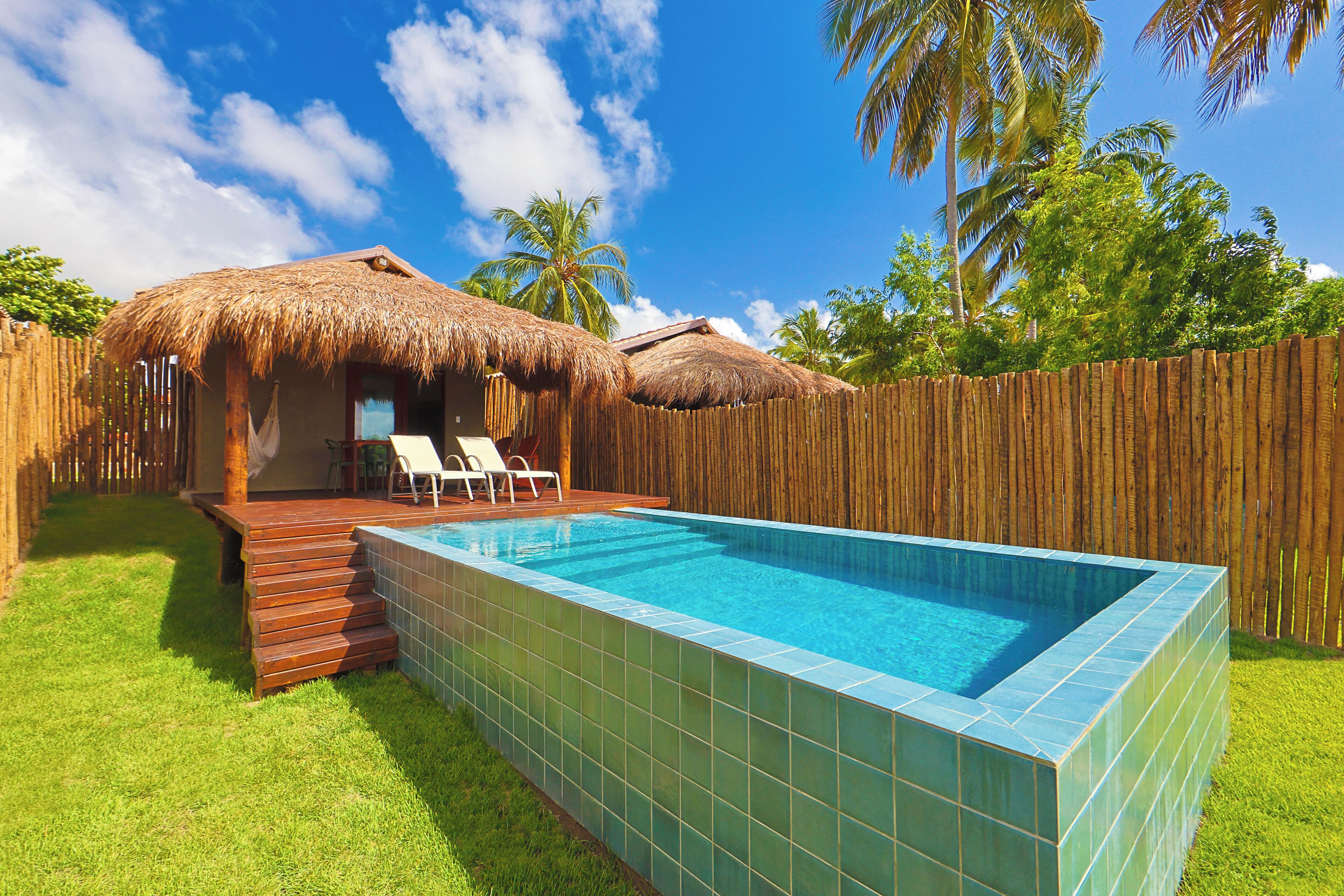 Ein Swimmingpool mit Holzterrasse und Liegestühlen, umgeben von einem Holzzaun und Palmen.