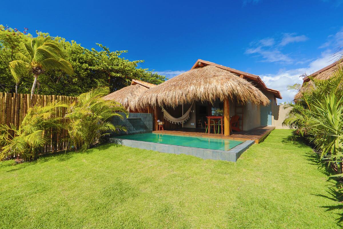 A thatched-roof house with a swimming pool, surrounded by lush greenery and a wooden fence.