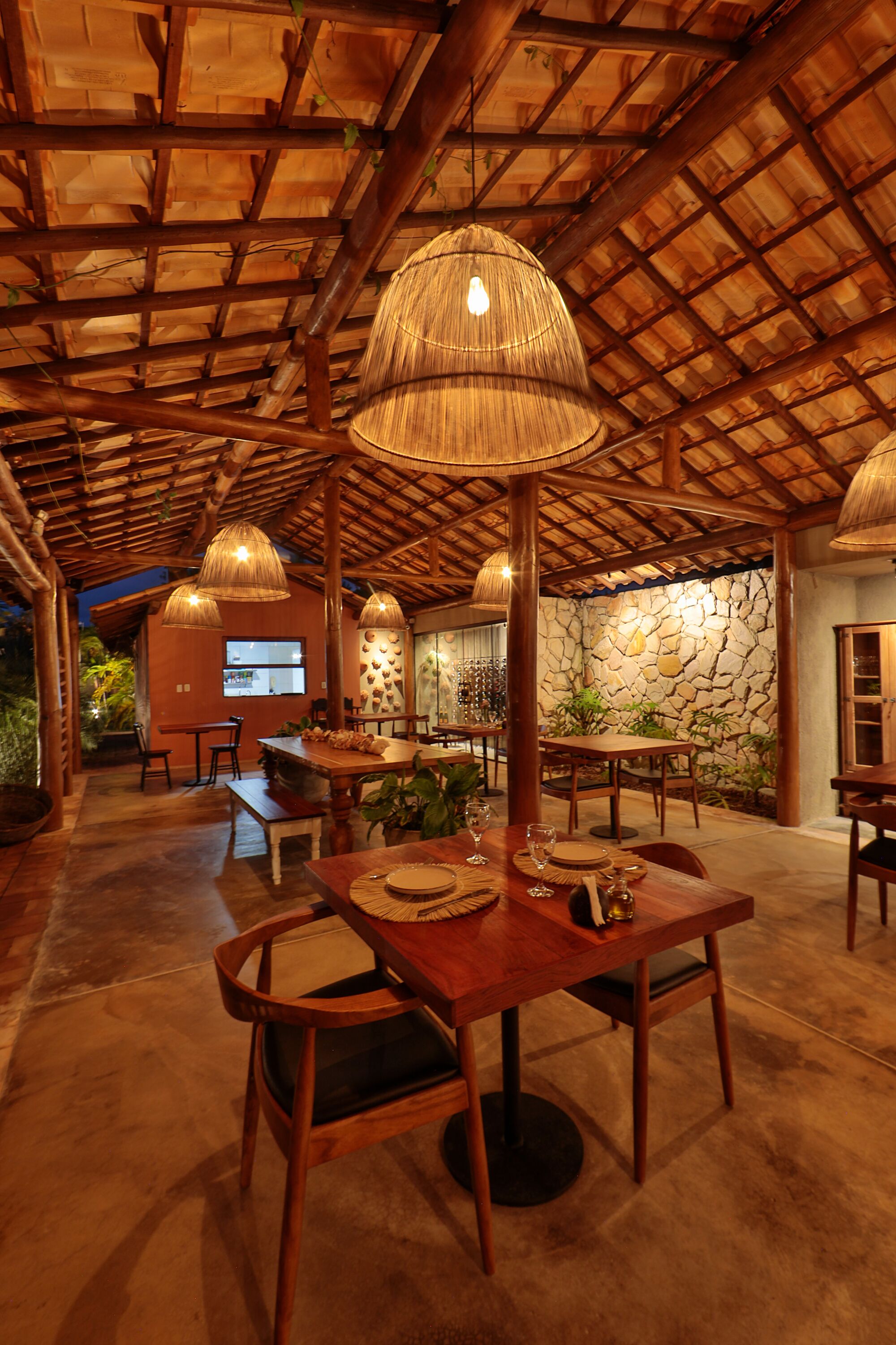 A restaurant with wooden tables and chairs, hanging lamps, and a stone wall.