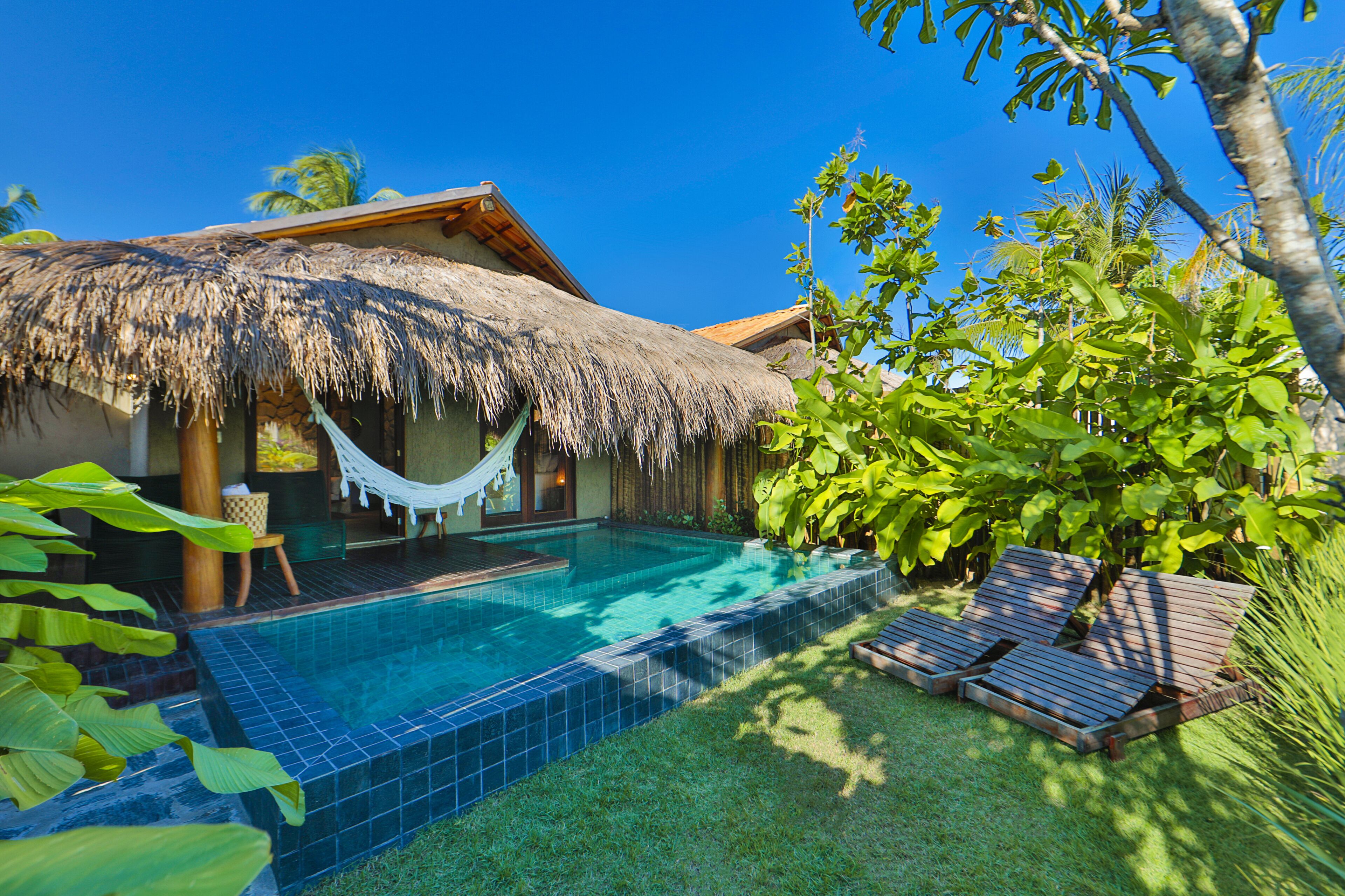 A pool area with a thatched roof, lounge chairs, and tropical plants.