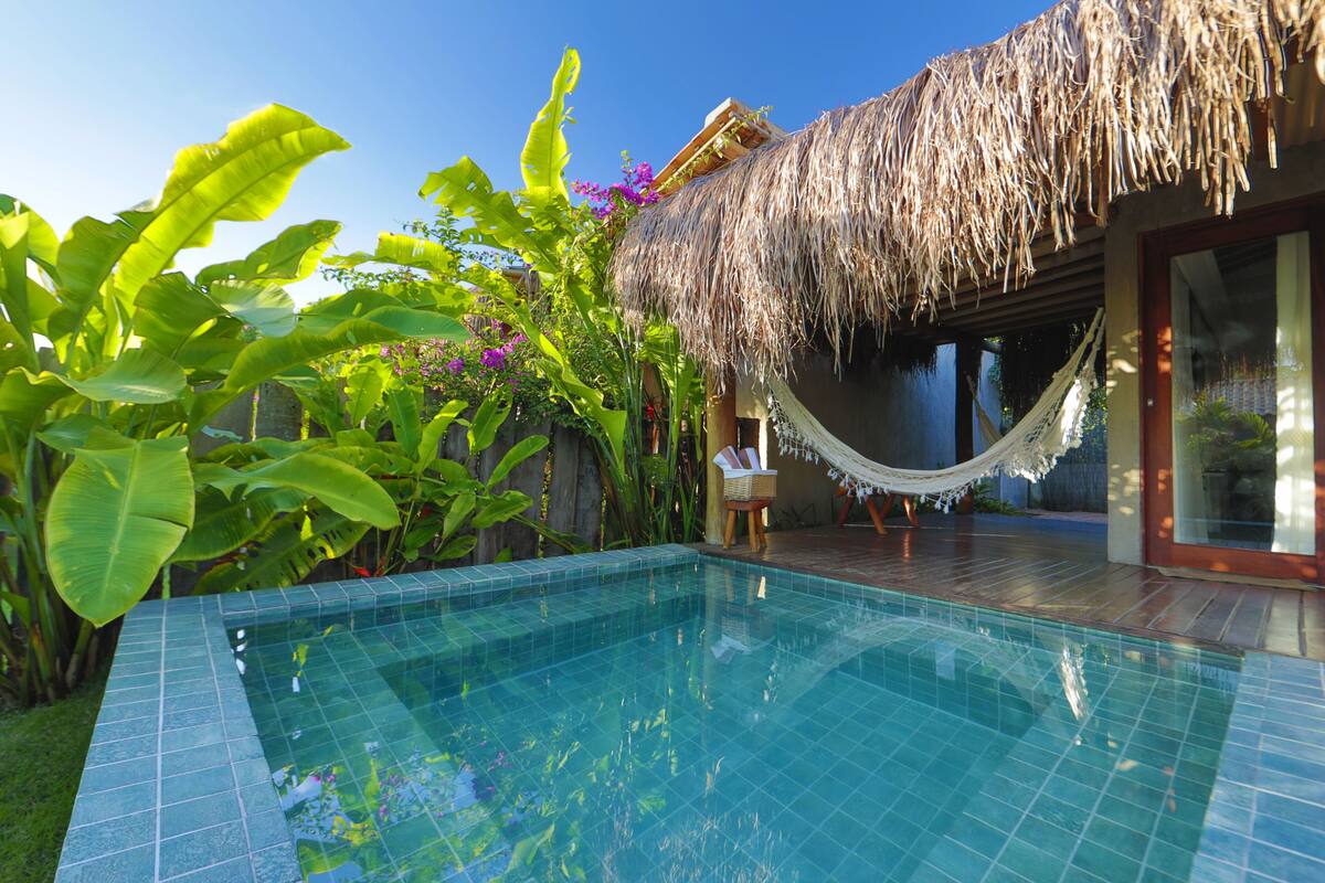 A swimming pool with a tiled edge, surrounded by lush greenery and a thatched-roof structure.