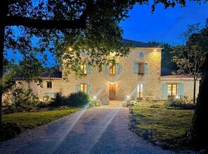 Un edificio de piedra con un camino de grava que lleva a una puerta de madera, rodeado de vegetación y árboles.