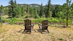 Two Adirondack chairs facing a stream with a forested backdrop and mountains in the distance.