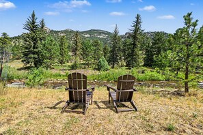 Two Adirondack chairs facing a stream with a forested backdrop and mountains in the distance.