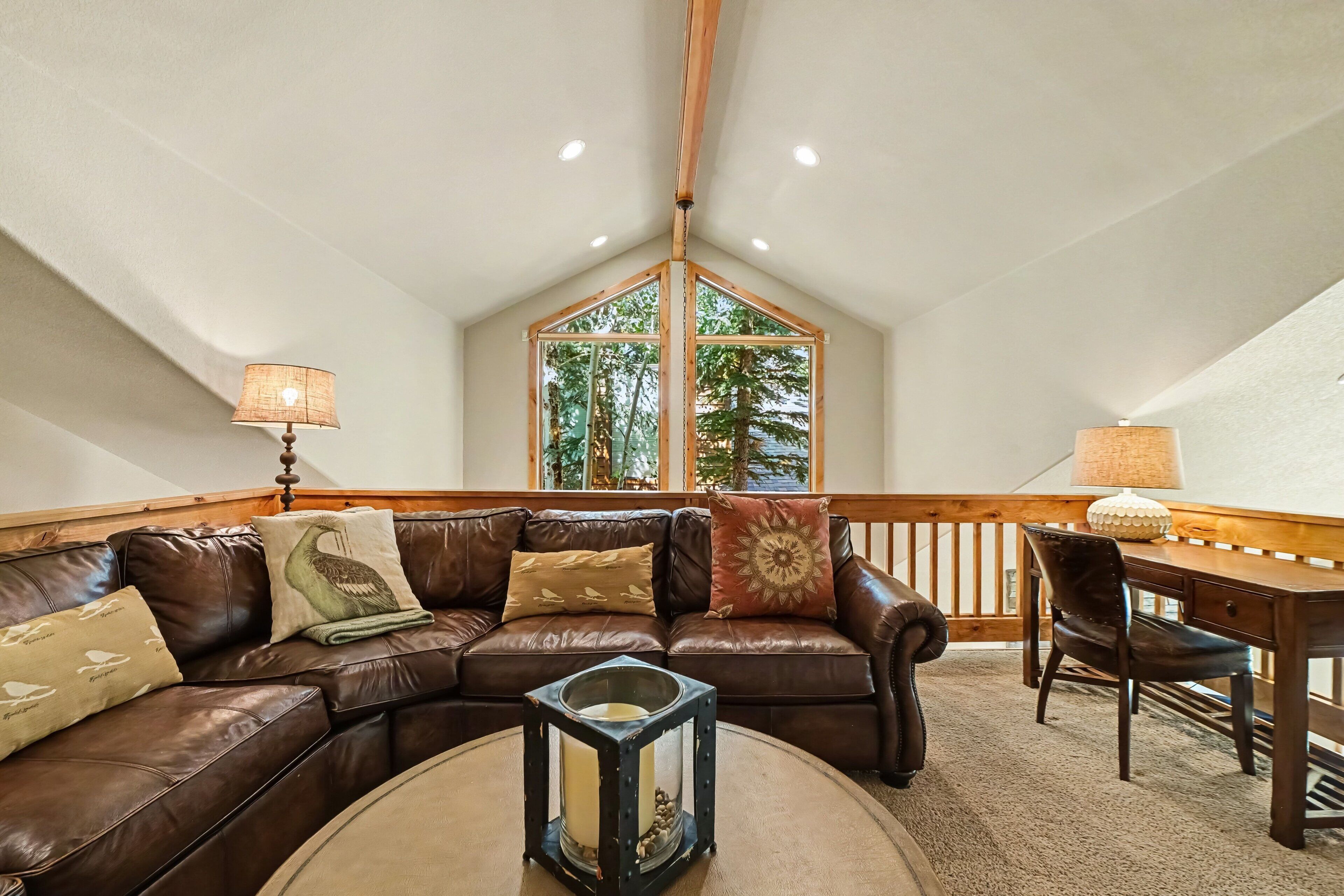 A cozy living room with a brown leather sofa, a round coffee table, and a wooden desk with a chair. A large window with a view of trees is visible.