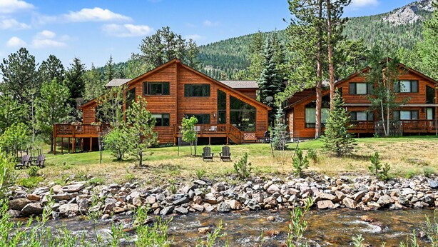 A lodge with multiple wooden cabins, a rocky stream in the foreground, and a mountainous backdrop.