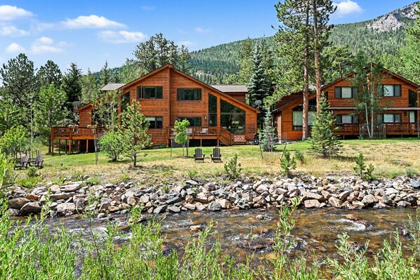 A lodge with multiple wooden cabins, a rocky stream in the foreground, and a mountainous backdrop.