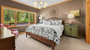 A bedroom with a large bed, a window with a view of greenery, a chandelier, and a green nightstand.