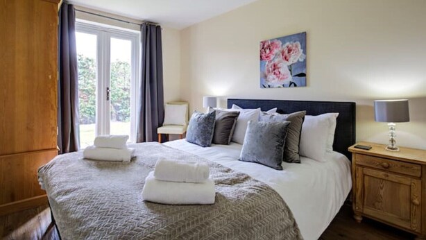 Cama bem arrumada com travesseiros e cobertor, mesa de cabeceira com abajur, quadro na parede e janela com cortinas.