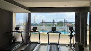 A gym with treadmills and a stationary bike, large windows with a view of a swimming pool and buildings. - La Santir by PSR (Pattaya)
