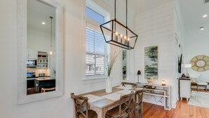 Een eetruimte met een houten tafel en stoelen, een hanglamp en een uitzicht op de keuken door een raam.