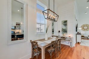 Een eetruimte met een houten tafel en stoelen, een hanglamp en een uitzicht op de keuken door een raam.