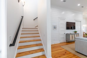 Un escalier avec des marches en bois et des rampes noires, menant à un intérieur blanc avec un canapé gris et un téléviseur fixé au-dessus d’un meuble.